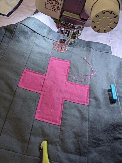 A quilt under the sewing machine head. It has a hot pink plus sign set in a grey background. The plus has been echoed on the inside in hot pink thread.