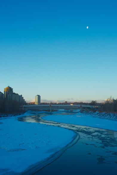 Partially frozen river at dusk. Dot of a half moon in the air.