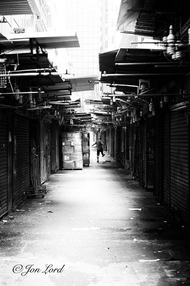 This is a dark and grungy black and white street photo in portrait format of a dim and dark alley with shuttered market stalls on either side and in the distance a man kicking an object. Jardine's Crescent, Hong Kong (2013).
The camera is located in the centre of this dim passageway, angled along it and about a metre and half above the ground. Extending away from the camera on both sides are closed market stalls, all with their shutters drawn down closed. The alley is about two and a half metres wide and stretches forward in a straight line for about fifty metres. Some of the market stalls have corrugated metal panels roughly placed above the entrances, all the stalls have small spiral neon lights hanging over the front. The rough concrete ground in front of the camera is brightly lit in the centre by the morning sun, with deep shadows on the side and is scattered with small pieces of litter. On the left there is an empty cloths rack a few metres in front of the camera and further on is a pallet of large cardboard boxes wrapped together with clear plastic. Perhaps ten metres from the camera and on the right side is the back of our man kicking out at some unknown object. Through the gaps in the corrugated roof panels is a bright sky, overexposed to brilliant white with the vague outline of a glass fronted tower block a hundred metres away.
