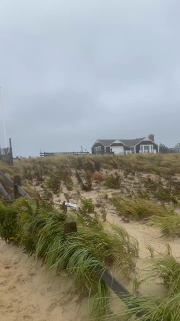 Video at the beach. east coast. Begins focused west on a red flag that is straight out from the North wind. Then pans to classic, shield beach homes with a grassy dune in front.  As the shot moves to the East - the rough surf and intense winds have eroded the beach