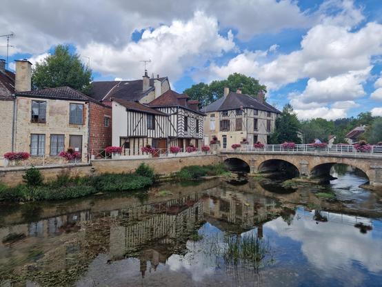 Village in the region Champagne in France. A river and three arches stone bridge and flowers in front (bridge goes from the right to the middle of the picture. Beuind it houses, some with "Fachwerk" beams. Blue sjy with large white clouds. The houses are mirrored in the water.