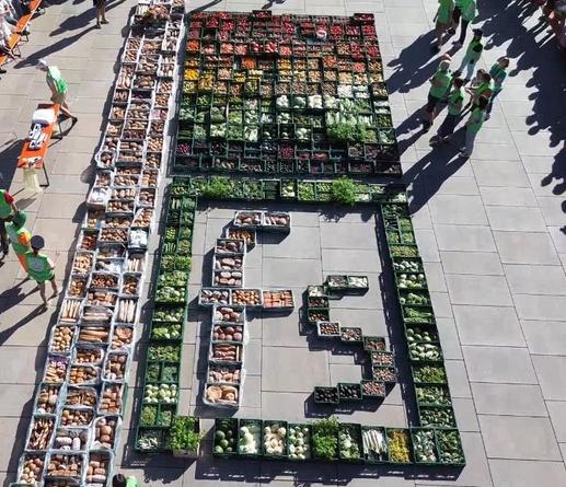 foodsharing am Tag der Lebenmittelrettung, Drohnenbild aus der Vogelperspektive auf 2.5 Tonnen Lebensmittel , Farblich sortiert in Kisten - die eine Regenbogenfahne und ein "fs" Symbol bilden - an einem sehr sonnigen Tag. Außen Stehen sehr viele Foodsharer herum