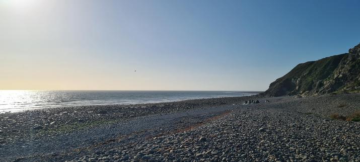 A grey pebble beach, with small cliffs to the right and the sea (which looks calmer than it really was) to the right. The sky is divided in two thanks to the sun, which is just out of the shot to the left: the sky on the left side is a soft peach while on the right, it's a hazy blue.
