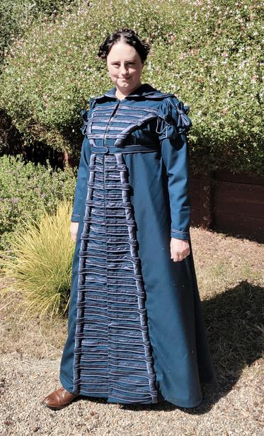 A blue coat dress covered in military style trim. The dress is high waisted, with an a-line skirt.

The dress is blue wool, trimmed with piped silk satin. It has a double collar, with the under side one so wide it touches the shoulders. The collar is bound in satin and pipped, because doing one or the other isn't enough.

There are 34 1 inch horizontal bands, from hem to neck, each side pipped. The outside edge of the bands is trimmed in a super fat piping, a 1 inch tube of stuffed silk satin.The horizontal bands twist over and under the satin tube. The trim spans shoulder to shoulder, gets narrow at the waist, only to flair again at the hem, like 2 triangles touching at the top.

The sleeves are long, but don't hit the knuckles as was the fashion of the time. Over the long sleeve is a fancy puff sleeve. The sleeve is slashed so you can see the sleeve underneath, with each edge bound in satin piping. The puff sleeve fabric is caught up in a large (also piped) bow, with the knot being 3 rows of piping. The shiny satin against the dull wool creates textural contrast. 

My hair is done up in ringlets at the side of my face, parted in the middle. The rest of my hair is in a bun at the middle back of my head.