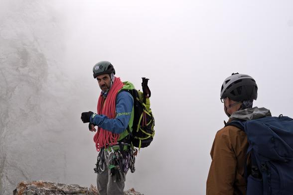 A person carrying a rope and standing near the edge of a cliff looks at the camera. Another person is looking away from the camera, at the first person. It's all grey cloud behind.