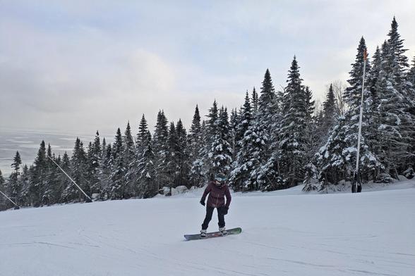 A person is on a snowboard on a fairly wide slope surrounded by snow-covered conifers. The Fleuve is visible in the background to the left.