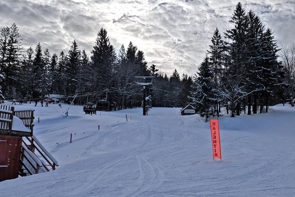 The base of the mountain at Mont Sutton. Mostly just a wide-open snow slope, with an orange warning banner to the right (RALENTIR backwards), and a chairlift in the middle of the picture. The sky is bright with broken cloud.