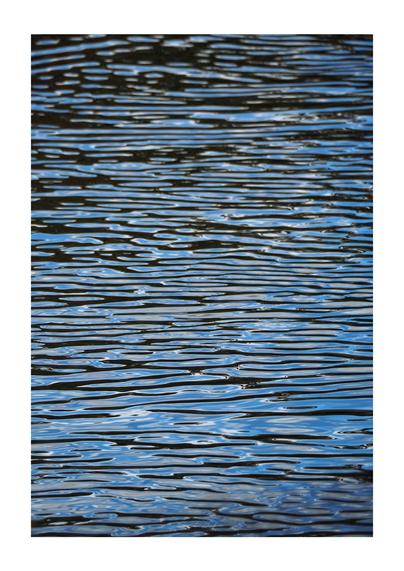 A portrait oriented photo, somewhat abstract in nature, of horizontal water ripples in black, grey, white and sky-blue. The image is in soft focus at the top and bottom of the photo.