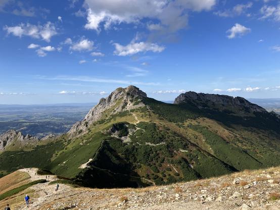View from the path up the mountain towards the back, where you can see another mountain (Giewont) with a characteristic shape of one narrow peak, then a break and then a wider ridge, with the narrow peak having a cross at the top. Behind it you can see some flat terrain in the distance with some villages.
