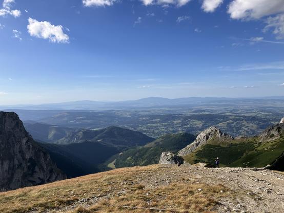 View from the path up the mountain, on a flat slope with yellowish grass, and the world below, with some hills, forests, villages, and some other mountains on the horizon. The sky is blue and you can see the landscape quite far from the mountains from this elevation.