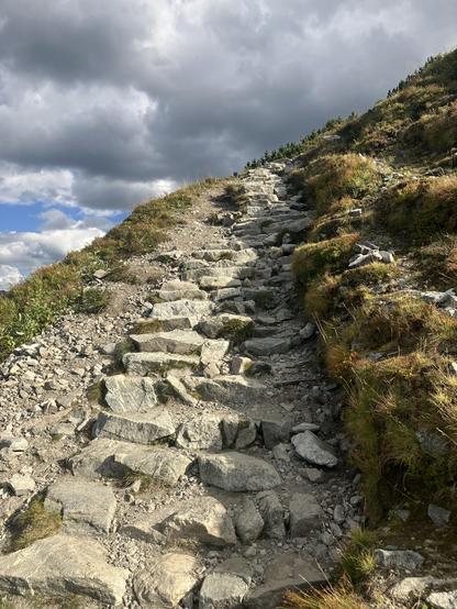 Close up on the stoney stairs up on this section of the trail, with yellow grass on the side and some quite dark gray clouds above.