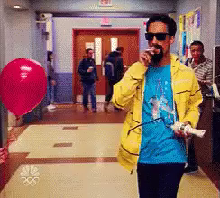Danny Pudi dressed as Snoop Dog walks down a school corridor and pops the red balloon that is being held by a kid with his cigarette or more likely joint