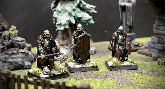 Three heavily armoured skeleton warriors adorned in reds on wintery bases stand in a green courtyard.