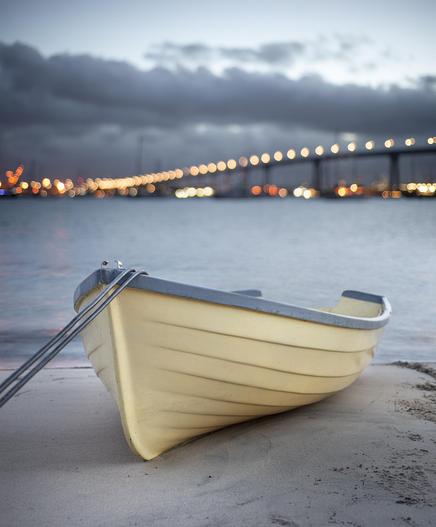 A color photograph of a small boat resting on a beach at Coronado, San Diego, California. Lights from the Coronado Bridge stretch across San Diego Bay in the background at dawn.