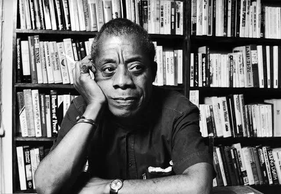 A black-and-white portrait of a man resting his head on one hand in front of a bookshelf, looking thoughtfully to the side. He sits at a desk with his arms crossed, wearing a dark shirt and wristwatch.