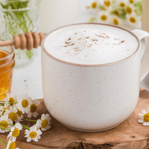 A white speckled ceramic mug filled with frothy coffee or latte topped with a dusting of cinnamon sits on a wooden slab, surrounded by small white daisies. A honey dipper, a jar of honey, and blurred flowers appear in the background.