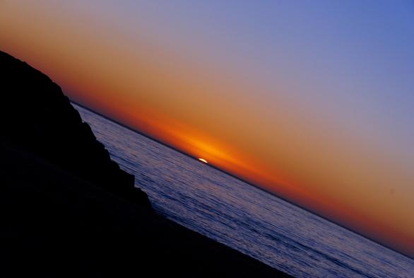 photo of the sun setting into the ocean.
the photo is on an angle, the horizon runs from the upper left corner to the lower right corner.
the sun, in the middle, is almost completely gone.  only the smallest sliver of it's disc is still visible.