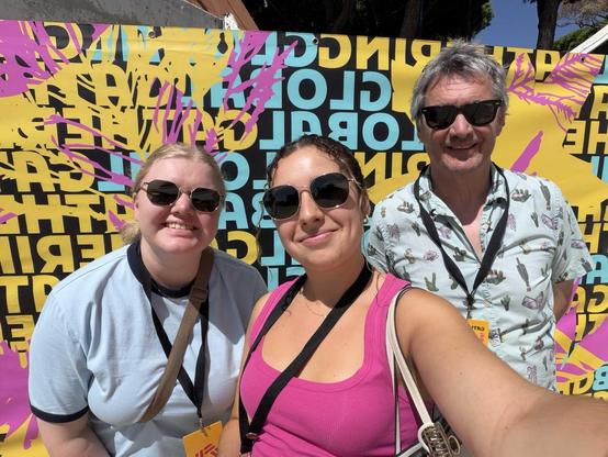 Saskia, Mariana, and Michael stand in front of a sign saying Global Gathering outside of the Global Gathering venue.