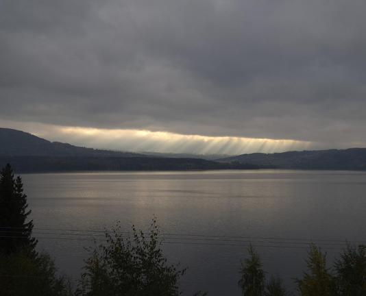 A thick blanket of clouds hangs over the hills on the other side of a lake; it is torn open like an elongated streak, through which the sun’s rays fall lengthwise. The water is still and dark.