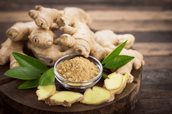 Fresh ginger roots arranged on a wooden board around a small glass bowl of ground ginger, with sliced ginger and green leaves in the foreground.