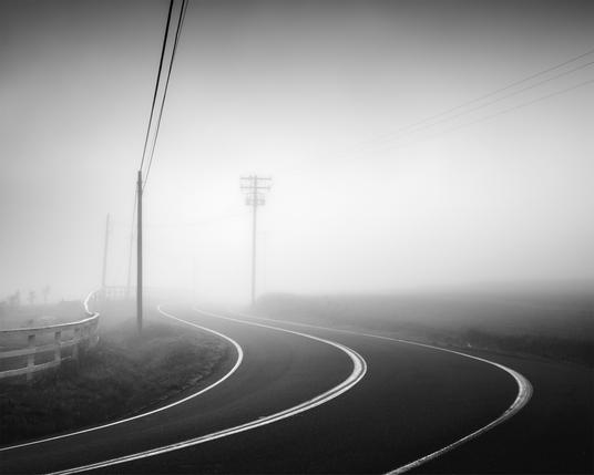 A black and white photograph of a curving road in East County San Diego, California. Heavy fog lights up in the background with sunrise.