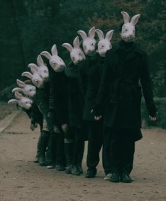 An old photo of people in all dark black and gray winter clothing, standing in a line on a wide dirt path in what appears to be a very green wooded area. 

The people are arranged by height, tallest in the front, shortest in the back. They are unnaturally close together and are all wearing big whole-head white rabbit masks. It’s a blankly staring cartoon rabbit sort of face. The frontmost and tallest person is standing up straight and with their arms out a little at the sides. Each person behind is leaning to their right side a little more than the one ahead of them, so the line of people creates a progressively leaning array of the white rabbit heads, the last few shortest people almost leaning fully sideways.

Some are looking at the front person a bit and some are facing more toward the camera.

The whole picture has a slight greenish cast and looks somewhat middle 20th Century. It’s meant to be weird, obviously, but is maybe unintentionally more spooky looking than even was planned.