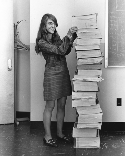 A black and white photo: A younger Margaret Hamilton standing next to the navigation software that she and her MIT team produced for the Apollo Project in 1969.