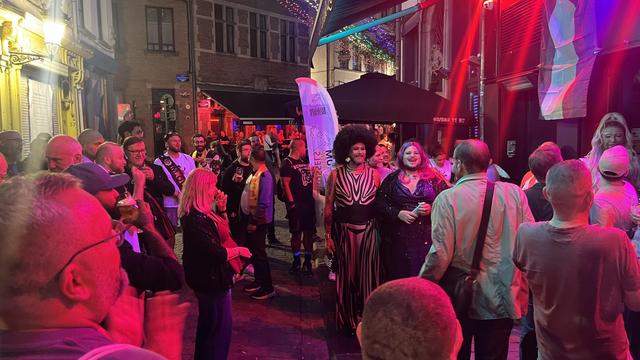 A crowd gathers on the street lit in red. There’s a banner for Belgium Bear Pride Week and two bearded drag queens smiling.