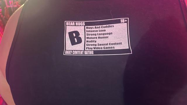 A label looks like a video game rating on the back of a bear’s tank-top: rated B for Bear Hugs. Hugs And Cuddles, Intense Love, Strong Language, Mature Humor, Nudity, Strong Sexual Content, Play Video Games.
