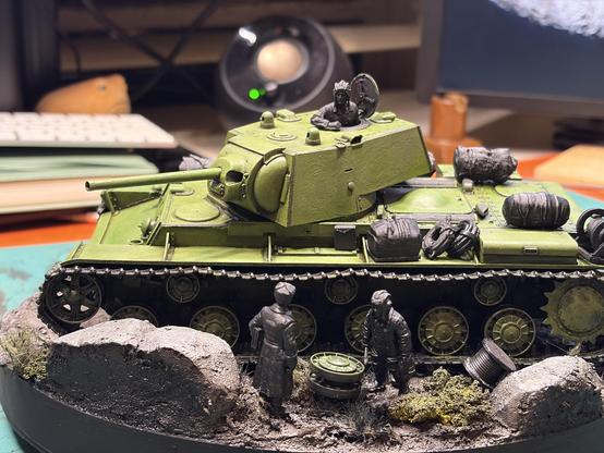 A scale model of a Soviet KV-1 heavy tank on a small diorama base. The tank is painted in bright weathered green. A tank crewman figure leans out of the open turret hatch. Two unpainted dark grey figures stand by the tracks, next to a spare road wheel and a cable spool.