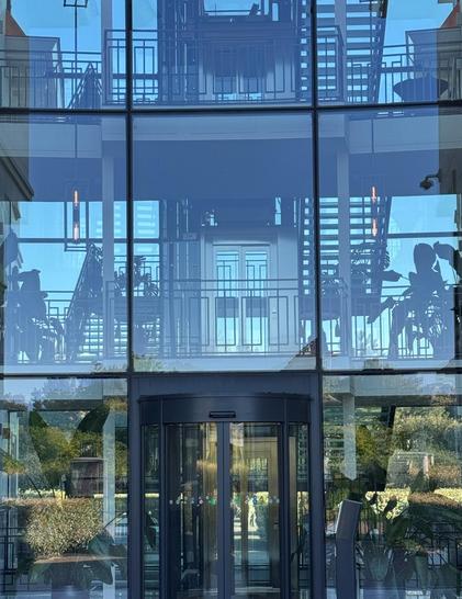A modern glass building featuring a revolving door. The reflections of exterior greenery and interior staircases are visible in the glass.
Ein modernes Glasgebäude mit einer Drehtür. Im Glas spiegeln sich die Begrünung im Außenbereich und die Treppen im Inneren wider.