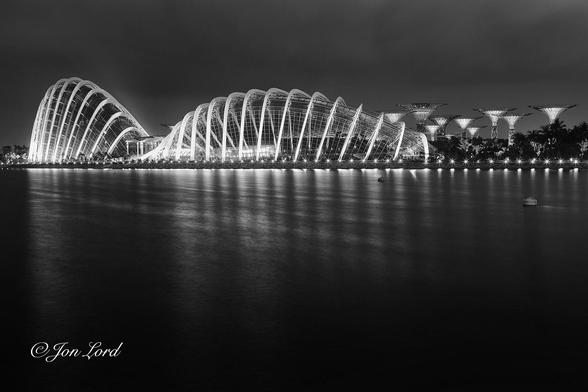 This is a black and white night time photo in landscape format of a body of smooth, dark water with two brightly lit futuristic giant glass buildings with a ten large tree like structures in the background. Gardens by the Bay, Singapore (2012).
Rising from the base of the photo to the two-thirds up level is a body of jet black water. The water is flat calm with light streak reflections from the buildings beyond. The only things on the water are two cylindrical buoys to the right of centre. The distant waterfront is perhaps one to two hundred metres away with lights evenly spaced along its length. The camera is located about two metres above the water and directed at the futuristic buildings on the opposite shore. The two giant buildings have a wave or serpentine shape to them or possibly that of a slug! The building directly ahead of the camera is the Flower Dome and is viewed in profile. The main structure appears to be fourteen or fifteen large white arches, the largest being in the centre and then sloping gently downwards towards each end. The arches support the glass roof structure and are brightly lit. To the right and slightly more distant are ten of the eighteen illuminated 'Supertrees' that have a tall cylindrical tubes supporting a wide inverted cone with a flat top. These vary in height from twenty-five to fifty metres. To the left of the Flower Dome is a further similar, but taller and more arched building. This is the Cloud Forrest with a similar arched structure supporting a glass covered interior. This building is viewed obliquely with one end and side in view. Above is a low, overcast sky with some parts of the clouds lit from the buildings below.