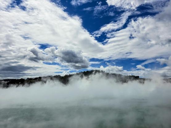 Steam is boiling off a pool of hot spring, clouds above, patches of sky, a glimpse of treetops