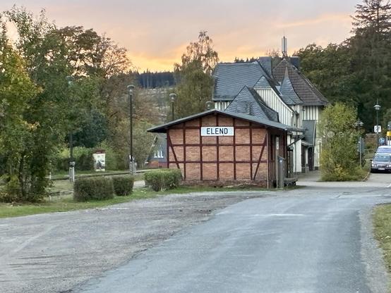 Ein Bahnhofsgebäude auf den der Ortsname „Elend“ steht. Dahinter ist der bewölkte Himmel orange eingefärbt