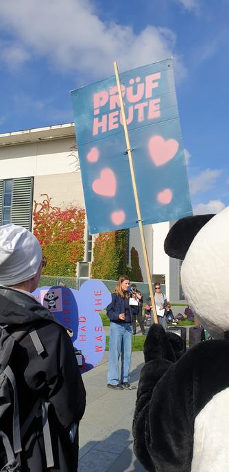 Luisa Neubauer spricht vor dem Kanzleramt

Vor Corinna steht noch der Berliner Prüf Panda und hält ein Schild hoch mit der Aufschrift 

Prüf heute