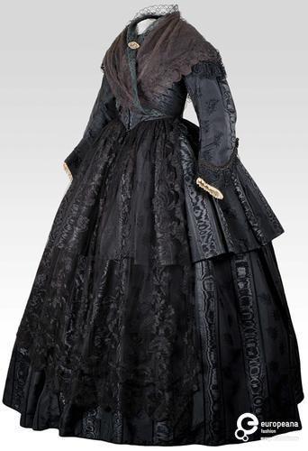 A black mid-Victorian gown in black moire silk, aka watered silk. The silk has been treated to the sheen ripples like water, in a very organic way. 

The gown is high necked, and has a shallow V at the center front. The bodice details are obscured by a black lace shawl being criss-crossed over the front and tied in the back. The sleeves are simple and have a 2 part design, so they follow the natural curve of the arm. There is additional texture at the cuff, but the photo doesn't have the resolution to figure out what. 

The long, dome shaped skirt has 2 tiers, with the top tier ending just above the knee. The skirt shows off the effect of the moire to a beautiful effect.