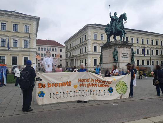 Menschen halten ein Banner mit der bunten Aufschrift: Es brennt! Hand in Hand für ein gutes Leben! Darunter eine Menschenkette aus Strichmännchen.
Dahinter day Reiterdenkmal eines bayrischen Königs, und eine große Figur, die die Omas gegen Rechts gebastelt haben.