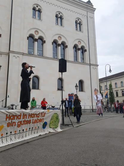 Die Kundgebungsbühne: Gerade sorgt Gündalein (eine junge Schwarze Frau in einem schwarzen Mantel) für Stimmung, vorne an der Bühne hängt wieder das Banner vom ersten Bild. Im Hintergrund stehen die Rednerinnen von GermanZero und Greenpeace sowie der Moderator bereit.