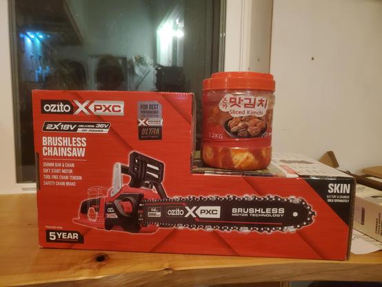 Box claiming to contain an Ozito battery chainsaw is on a wooden bench. On top of it is a jar claiming to contain Kimchi. The jar is mostly transparent and what's inside seems to match the claim but we have no such visibility inside the box.