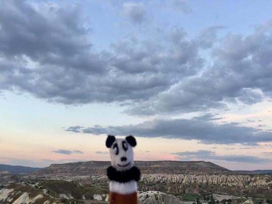 A small black-and-white puppet, called Panda, stands in the foreground, overlooking the rocky landscape of Göreme at sunset. The sky is filled with dramatic blue-grey clouds, with pale pink and yellow light near the horizon. Photo is by Panda Mery, gizmonaut.net.