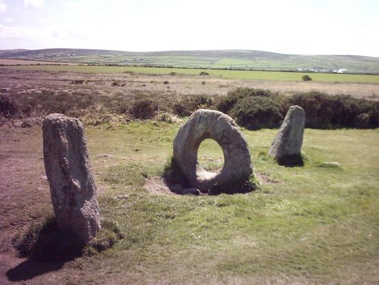 Colour photo of the Mên-an-Tol formation of standing stones in West Cornwall. The low, rolling moors, with fields on the lower slopes, can be seen in the distance beneath a pale blue sky with a few fluffy low clouds at either side, the hidden sun obliterating all details at the centre. The small stones of a low cairn or platform can be seen just to the left of the holed stone. All three stones have short shadows.