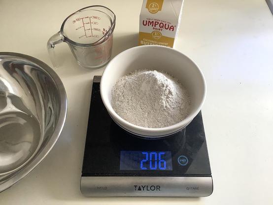 A bowl of rye flour on a kitchen scale and a small carton of buttermilk.