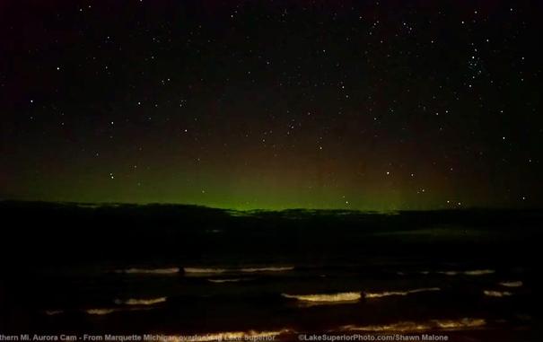 Greek bands of northern lights with a layer faint red above them stretch along a cloudy horizon. Waves from Lake Superior wash ashore in white lines. The dark sky is loaded with stars.