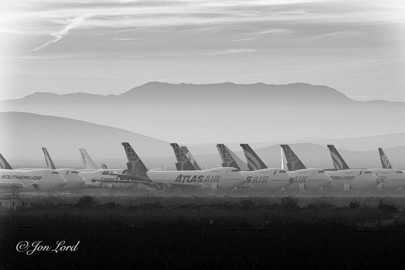 This is a black and white dawn photo in landscape format of a large number of parked Boeing 747 aircraft arranged in rows in longterm storage with a mountain range in the background. Mojave Air and Space Port, California (2016).
In the foreground and rising up to a little less than the quarter way up level is desert brush mostly deep in shadow. Above are the neat rows and lines of those parked Boeing 747 aircraft. They are view obliquely with the noses and right side in view and observed from a few hundred metres away. All the planes have their noses pointing to the right. The closest aircraft have a white fuselage and dark tail with the words 'Atlas Air' in large lettering in the centre of the fuselage. In the row behind, the aircraft again have similar colouring but with the words 'Southern Air' in large dark lettering. Most have their forward cargo doors open. Many have engines and other parts removed. In the background the air is hazy with a distant tall and long mountain range stretching across the photo from left to right. Above is a partly cloudy sky with only thin wisps of cirrus high clouds obscuring the heavens.