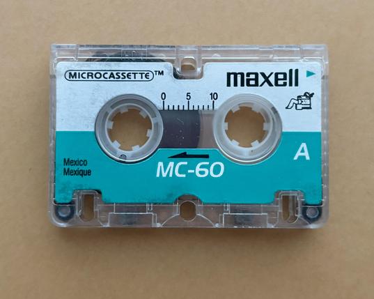 straight-on photo of a maxell mc-60 microcassette, made in mexico which may help date it. the upper half of the label is white and contains the maxell branding, the microcassette branding, the indicator showing where the write protect tab is, and the tape progress indicator. the lower half is teal and contains the country of origin, the a-side indication, the mc-60 branding, and the play direction indicator. in the upper half, below the maxell logotype is a tiny illustration of a man sitting in a chair, being blown away; a callback to a highly successful ad campaign (well, successful except that everyone seems to confuse it with the memorex ‘is it live?’ campaign…)
