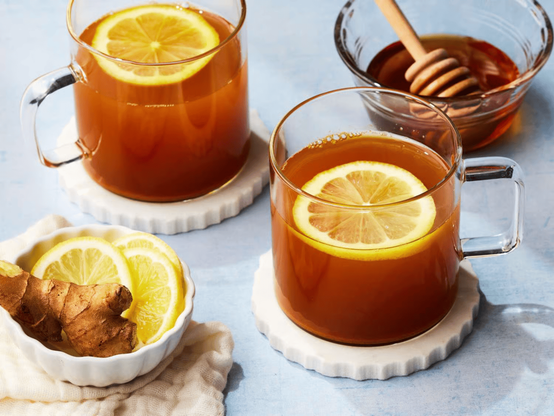 Two clear glass mugs of amber tea topped with lemon slices sit on white coasters, with a bowl of honey and a honey dipper in the background. A small dish of sliced lemon and fresh ginger rests at the lower left.
