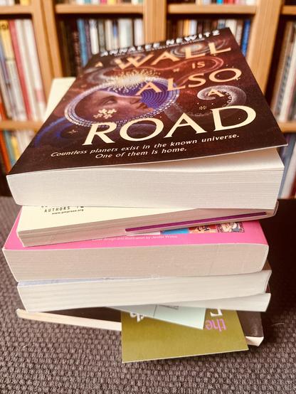 a bookstore-fresh paperback copy of annalee newitz's "a wall is also a road," atop a pile of other books, in front of a wall of still other books