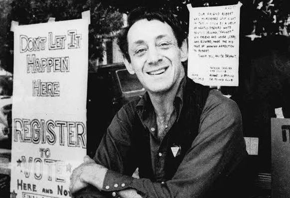 A black-and-white photo shows a smiling man seated with his arms crossed in front of signs; the large poster at left reads “DON’T LET IT HAPPEN HERE REGISTER TO VOTE,” with “HERE AND NOW” at the bottom. A smaller handwritten sign hangs behind him, but most of that text is hard to read.