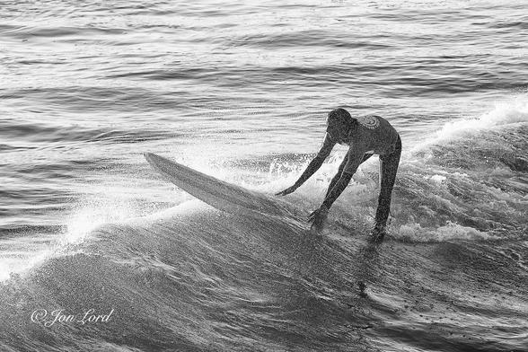 This is a black and white water sports photo in landscape format of a female teen, surfing shortly after sunrise on the crest of a breaking wave and leaning forward to maintain her balance. Hermosa Beach, California (2013).
The breaking Pacific wave stretches diagonally across the photo from the lower left corner, through the centre, upwards towards the upper right corner and is in motion from right to left. The sea occupies the entire frame but is divided between the calm and rippled water ahead of the wave in the upper left and the wave filling the lower right corner. The air around the wave is saturated with a storm of fine water droplets thrown up by the wave. To the right of centre is our teen surfer. She is viewed obliquely with her front and left side in view and in motion from right to left. The camera is located three metres away from the surfer and two metres above the water. Our surfer is partially obscured by the spray from the wave. She is wearing a t-shirt over a black wetsuit, has long dark hair and is bending forward with her hands holding the edges of her board. The forward third of the surfboard is ahead of the wave and hanging over the crest. The aft part of the board and surfer are just behind the crest.