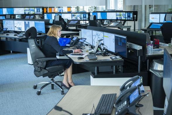 Zakelijk geklede vrouw op grote bureaustoel met voor haar op tafel 4 schermen. Zij zit midden in een kantoor met overal setjes van 4 of 5 schermen. Verder geen mensen te zien.